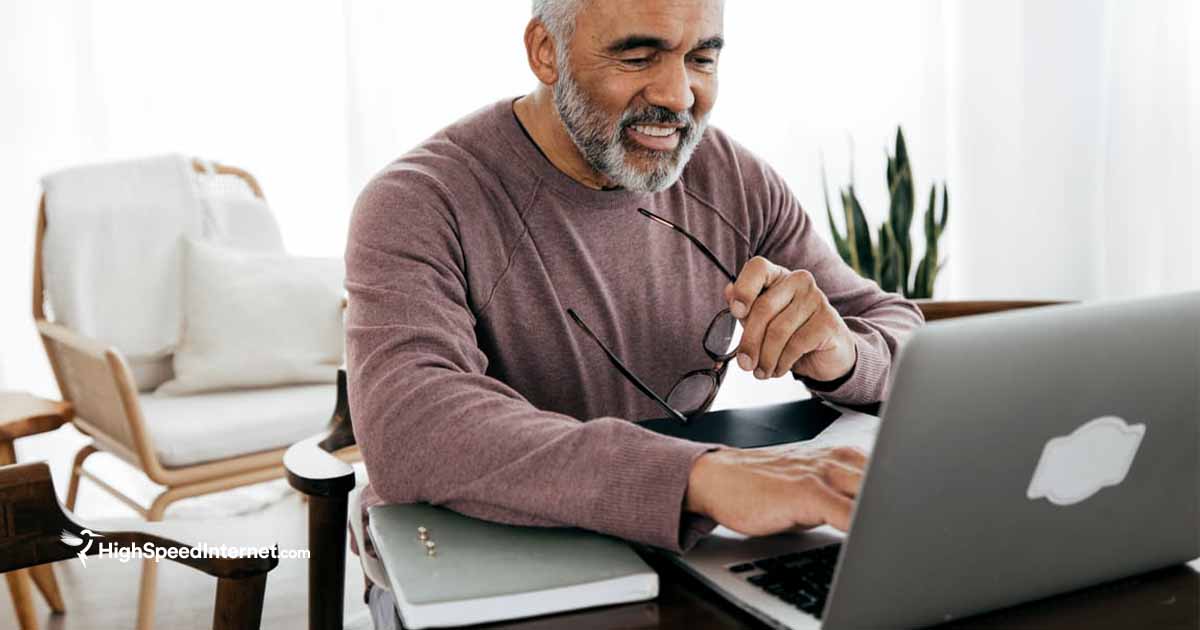 happy male sitting at desk using laptop while holding glasses in hand